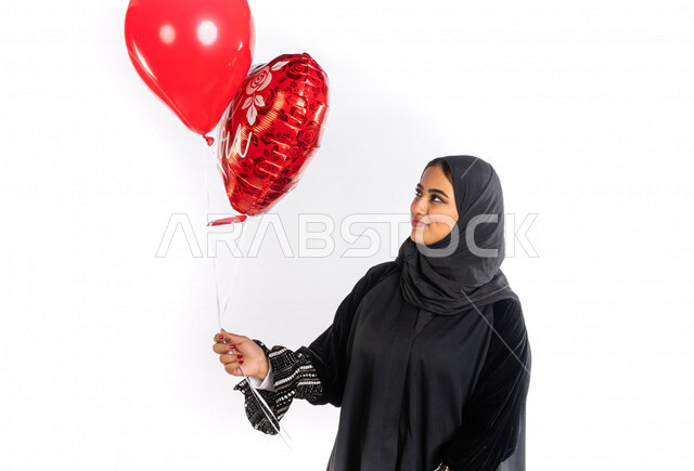 بورتريه لمرأة عربية خليجية سعودية تحمل بالونين هيليوم أحمر ، بإيماءات وجه ويدين مختلفة تدل على الحب والفرح والسعادة ، هدية بمناسبة يوم الأم ، هدية عيد الحب ، أعياد ومناسبات ، خلفية بيضاء ، مناسبات سعيدة ، مفاجأة ، عيد الميلاد