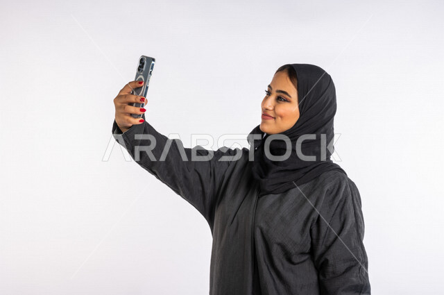 بورتريه لمرأة عربية خليجية سعودية ، تقوم بإلتقاط صور سيلفي تذكاريه بإستخدام الهاتف المحمول ، التسليه والترفيه ، إستخدام أجهزة تقنية حديثة ، إستخدام وسائل التواصل الإجتماعي ، إجراء مكالمة فيديو ، بث مباشر ، التواصل مع الأصدقاء ، خلفية بيضاء