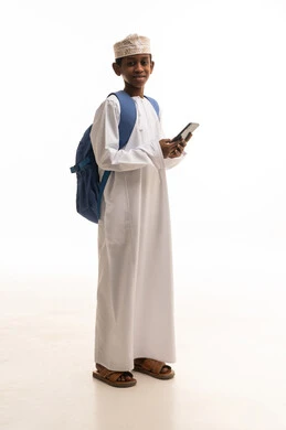 طفل عماني يرتدي الزي التقليدي مع حقيبة ظهر وهاتف ذكي
