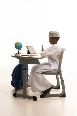 طالب عُماني يجلس خلف مكتب دراسي يستخدم جهازاً لوحياً طالب عُماني يجلس خلف مكتب دراسي يستخدم جهازاً لوحياً