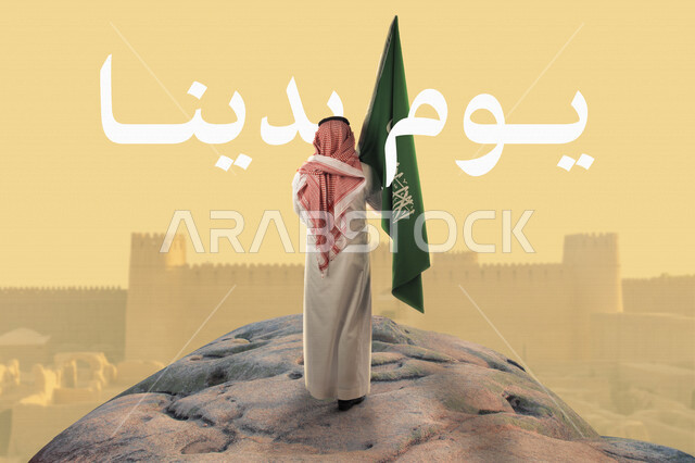 صورة من الخلف لرجل عربي خليجي سعودي يحمل علم السعودية وامامه المباني الأثرية التاريخية في السعودية، الدرعية القديمة في مدينة الرياض، ذكرى تأسيس الدولة السعودية، شعار يوم التأسيس، يوم بدينا