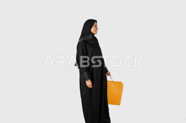 بورتريه بنت خليجية سعودية محجبة تحمل أكياس التسوق من المتجر , حقائب ورقية , ماركات عالمية , التسوق في المول , متعة التسوق في السعودية , خلفية بيضاء .