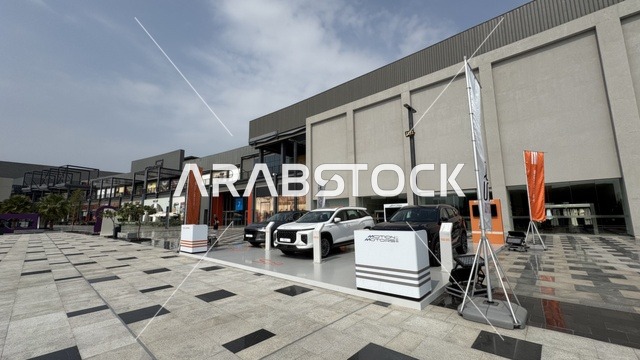 Parking in the outdoor parking lot of the U Walk complex in Jeddah, Saudi Arabia, an open walkway with a modern design, international and local restaurants and cafes, a comprehensive urban destination for walking, entertainment, and shopping, diverse brands, the exterior design of the Sinomi Mall building, seasonal events and recreational activities, outdoor seating and social spaces, vibrant family and youth atmosphere.