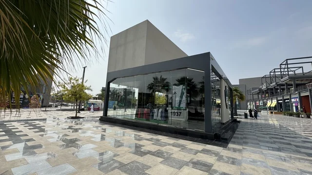 The Saudi Al-Ahli Club at the U Walk complex in Jeddah, Kingdom of Saudi Arabia, features an open walkway with a modern design, international and local restaurants and cafes, a comprehensive urban destination for walking, entertainment, and shopping, diverse brand names, the exterior design of the Sinomi Mall building, seasonal events and recreational activities, outdoor seating and social spaces, and a vibrant family and youth atmosphere.