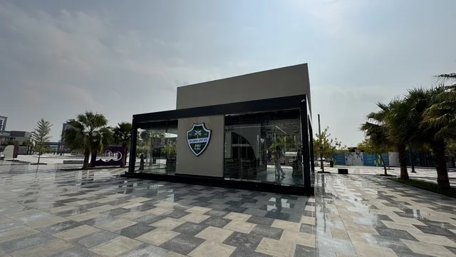 The Saudi Al-Ahli Club at the U Walk complex in Jeddah, Kingdom of Saudi Arabia, features an open walkway with a modern design, international and local restaurants and cafes, a comprehensive urban destination for walking, entertainment, and shopping, diverse brand names, the exterior design of the Sinomi Mall building, seasonal events and recreational activities, outdoor seating and social spaces, and a vibrant family and youth atmosphere.
