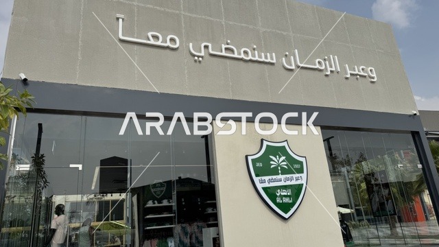 The Saudi Al-Ahli Club at the U Walk complex in Jeddah, Kingdom of Saudi Arabia, features an open walkway with a modern design, international and local restaurants and cafes, a comprehensive urban destination for walking, entertainment, and shopping, diverse brand names, the exterior design of the Sinomi Mall building, seasonal events and recreational activities, outdoor seating and social spaces, and a vibrant family and youth atmosphere.