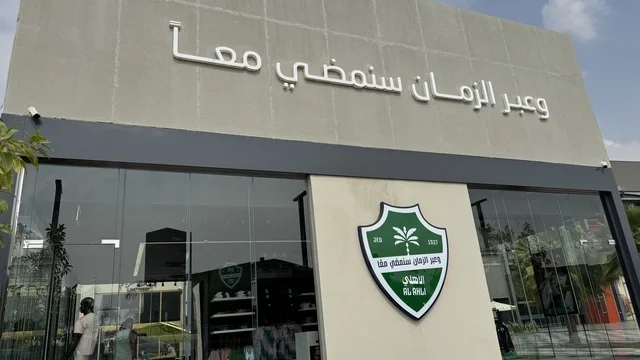 The Saudi Al-Ahli Club at the U Walk complex in Jeddah, Kingdom of Saudi Arabia, features an open walkway with a modern design, international and local restaurants and cafes, a comprehensive urban destination for walking, entertainment, and shopping, diverse brand names, the exterior design of the Sinomi Mall building, seasonal events and recreational activities, outdoor seating and social spaces, and a vibrant family and youth atmosphere.