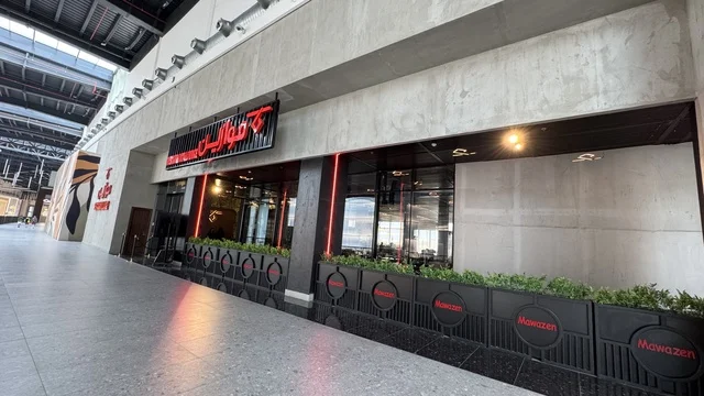 A brand at the U Walk complex in Jeddah, Saudi Arabia, February 15, 2024, an open walkway with a modern design, international and local restaurants and cafes, the exterior design of the Sinomi Mall building, a comprehensive urban destination for walking, entertainment, and shopping, outdoor seating and social spaces, a vibrant family and youth atmosphere.