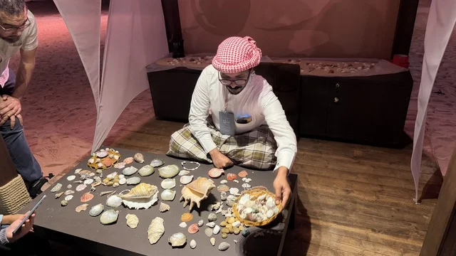 A man selling shells, displays of traditional shipbuilding stages and the tools and materials used, the wooden ships festival on the Al-Hamra beach in Jeddah, Saudi Arabia from January 12 to 16, 2026, maritime heritage, interactive activities, and cultural performances.