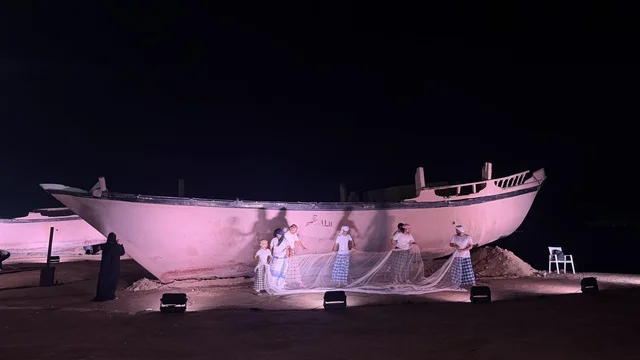 Next to a ship, a person wearing traditional attire plays a musical instrument, showcasing the stages of traditional shipbuilding, tools, and materials used. The Wooden Ships Festival on the Al-Hamra Beach in Jeddah, Saudi Arabia, from January 12 to 16, 2026, highlights maritime heritage, interactive activities, and cultural performances.