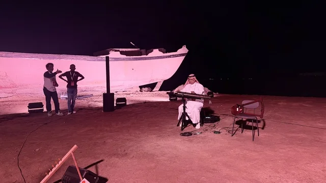 Next to a ship, a person wearing traditional attire plays a musical instrument, showcasing the stages of traditional shipbuilding, tools, and materials used. The Wooden Ships Festival on the Al-Hamra Beach in Jeddah, Saudi Arabia, from January 12 to 16, 2026, highlights maritime heritage, interactive activities, and cultural performances.