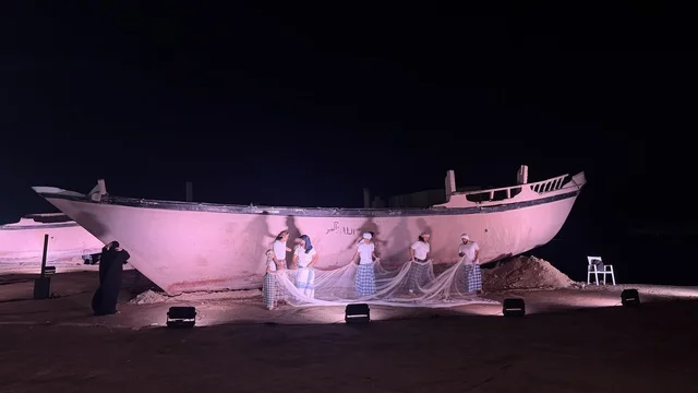 Next to a ship, a person wearing traditional attire plays a musical instrument, showcasing the stages of traditional shipbuilding, tools, and materials used. The Wooden Ships Festival on the Al-Hamra Beach in Jeddah, Saudi Arabia, from January 12 to 16, 2026, highlights maritime heritage, interactive activities, and cultural performances.