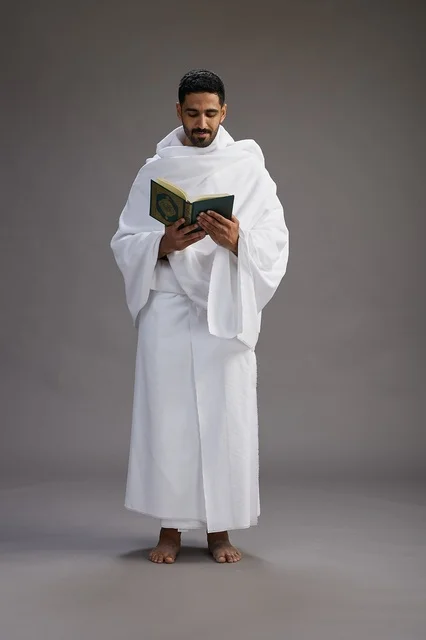A portrait of a Saudi man in Ihram clothing reading the Quran during Hajj, full-body, gray background.