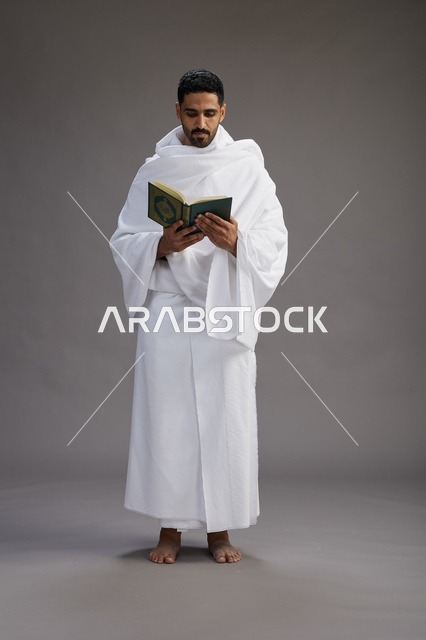 A portrait of a Saudi man in Ihram clothing reading the Quran during Hajj, full-body, gray background.