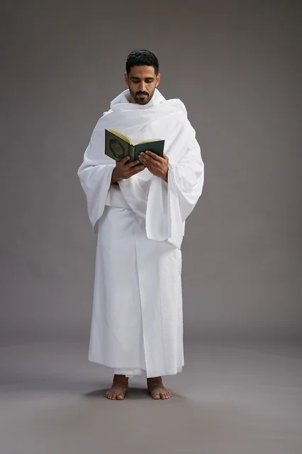 A portrait of a Saudi man in Ihram clothing reading the Quran during Hajj, full-body, gray background.
