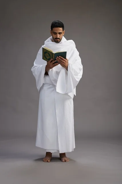 A portrait of a Saudi man in Ihram clothing reading the Quran during Hajj, full-body, gray background.