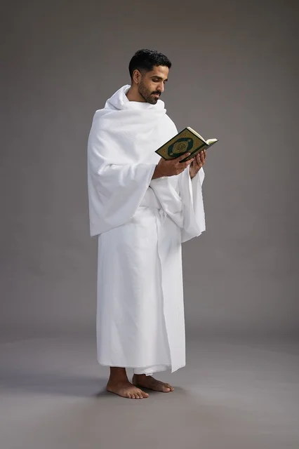 A portrait of a Saudi man in Ihram clothing reading the Quran during Hajj, full-body, gray background.