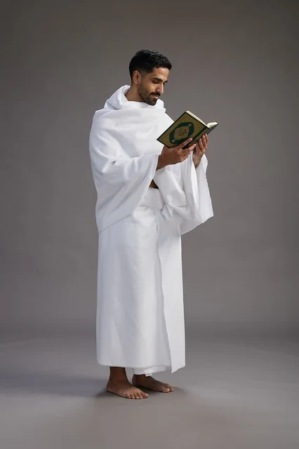 A portrait of a Saudi man in Ihram clothing reading the Quran during Hajj, full-body, gray background.