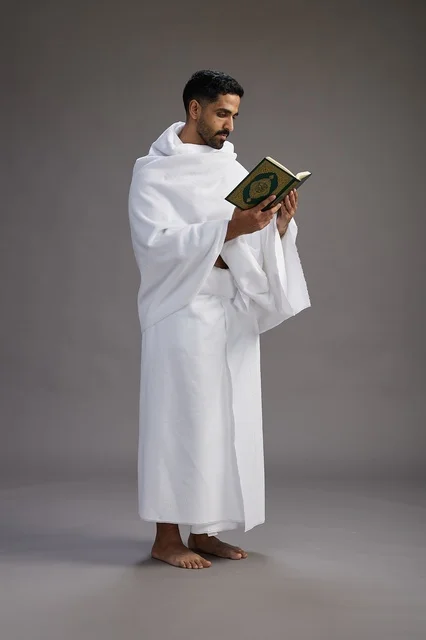 A portrait of a Saudi man in Ihram clothing reading the Quran during Hajj, full-body, gray background.