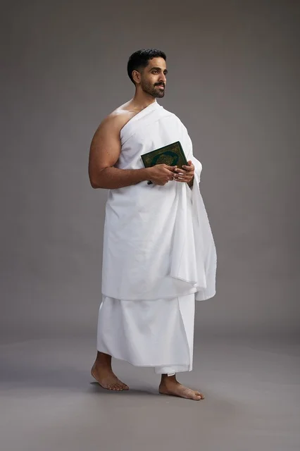 A portrait of a Saudi man in Ihram clothing reading the Quran during Hajj, full-body, gray background.