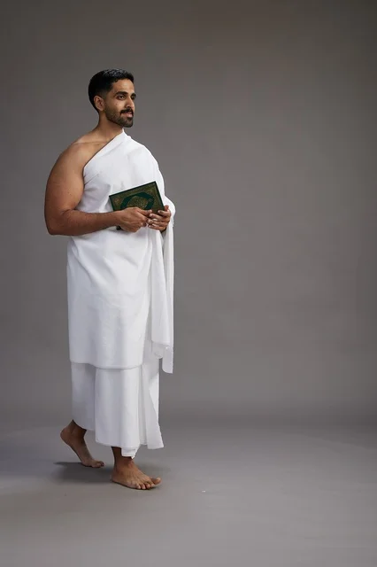 A portrait of a Saudi man in Ihram clothing reading the Quran during Hajj, full-body, gray background.