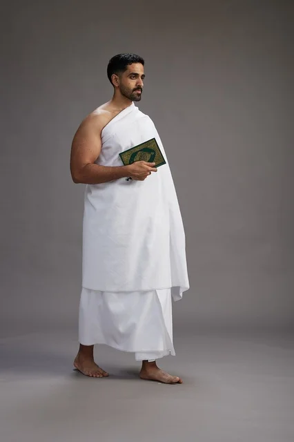 A portrait of a Saudi man in Ihram clothing reading the Quran during Hajj, full-body, gray background.
