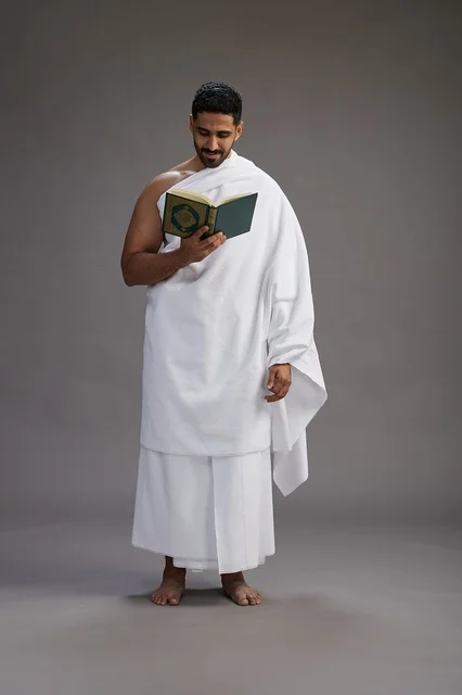 A portrait of a Saudi man in Ihram clothing reading the Quran during Hajj, full-body, gray background.