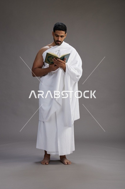 A portrait of a Saudi man in Ihram clothing reading the Quran during Hajj, full-body, gray background.