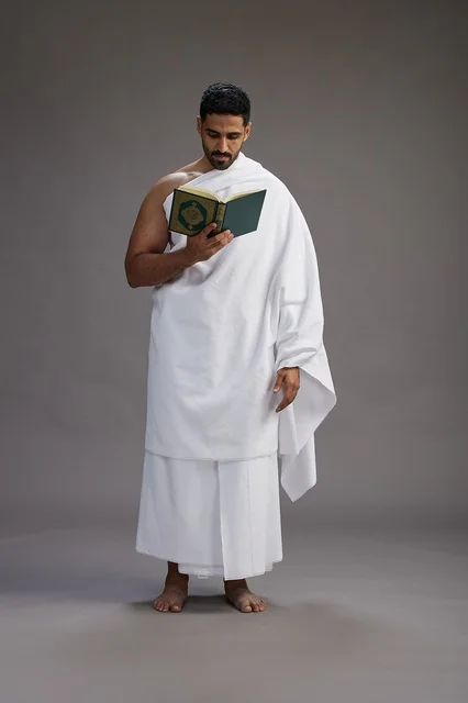 A portrait of a Saudi man in Ihram clothing reading the Quran during Hajj, full-body, gray background.