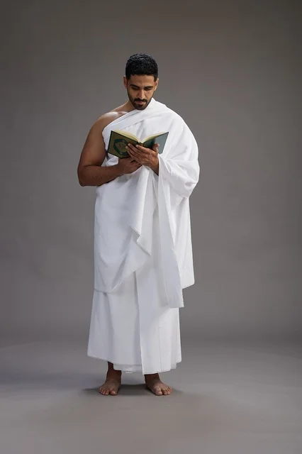 A portrait of a Saudi man in Ihram clothing reading the Quran during Hajj, full-body, gray background.
