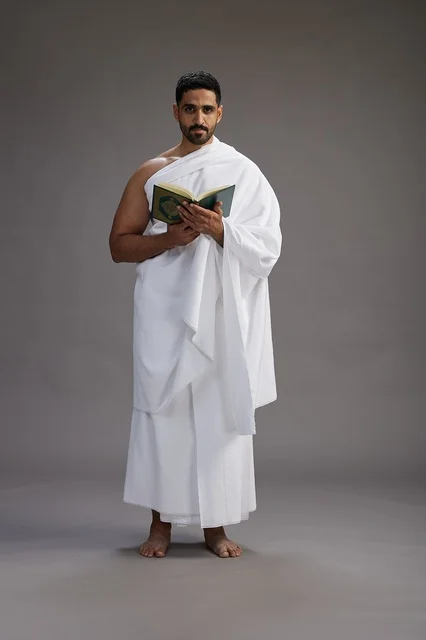 A portrait of a Saudi man in Ihram clothing reading the Quran during Hajj, full-body, gray background.