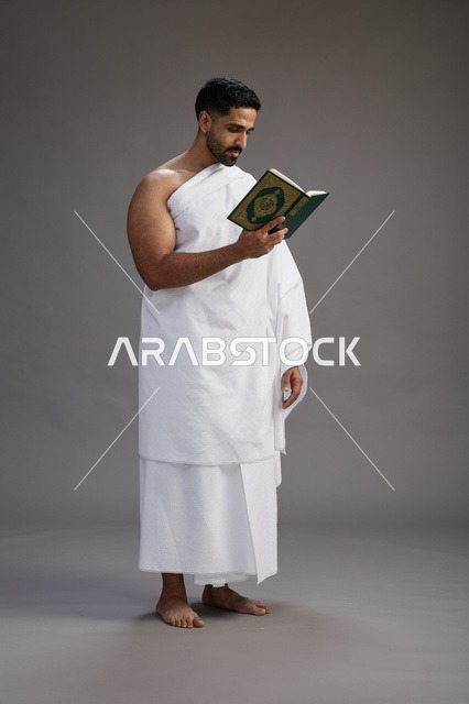 A portrait of a Saudi man in Ihram clothing reading the Quran during Hajj, full-body, gray background.