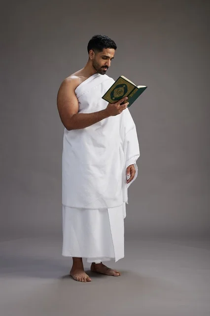 A portrait of a Saudi man in Ihram clothing reading the Quran during Hajj, full-body, gray background.