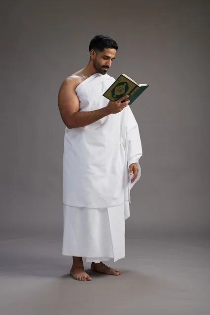 A portrait of a Saudi man in Ihram clothing reading the Quran during Hajj, full-body, gray background.