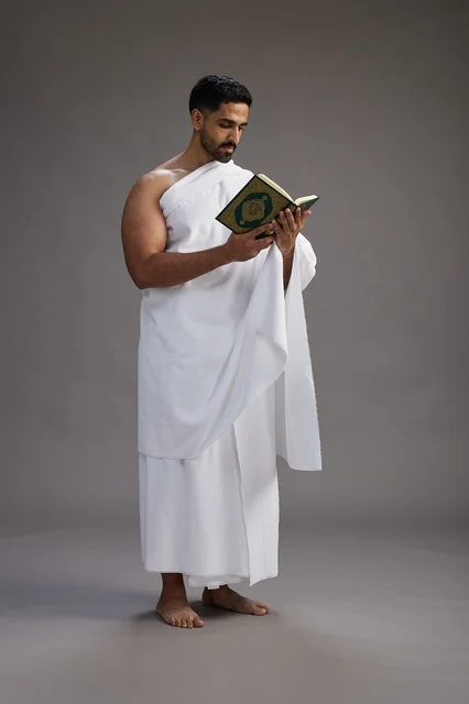 A portrait of a Saudi man in Ihram clothing reading the Quran during Hajj, full-body, gray background.