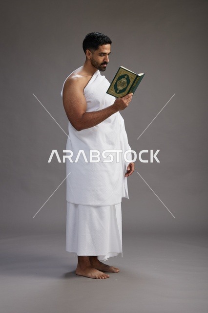 A portrait of a Saudi man in Ihram clothing reading the Quran during Hajj, full-body, gray background.