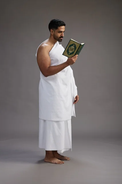 A portrait of a Saudi man in Ihram clothing reading the Quran during Hajj, full-body, gray background.