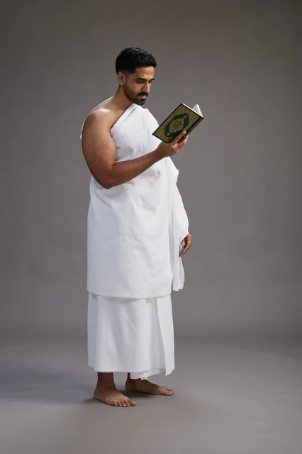A portrait of a Saudi man in Ihram clothing reading the Quran during Hajj, full-body, gray background.