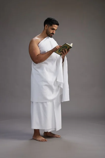 A portrait of a Saudi man in Ihram clothing reading the Quran during Hajj, full-body, gray background.
