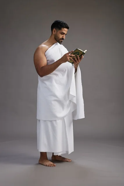 A portrait of a Saudi man in Ihram clothing reading the Quran during Hajj, full-body, gray background.