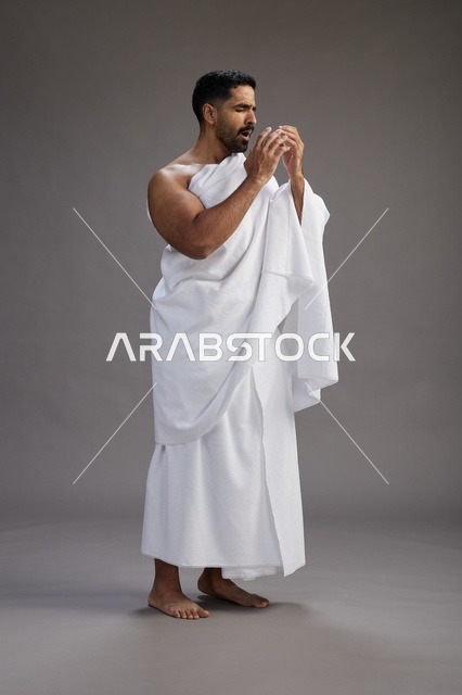 بورتريه لرجل سعودي بملابس الإحرام يعطس ويغطي فمه باحترام, مفهوم الحج والعمره, الجسم بالطول الكامل, خلفيه رماديه
