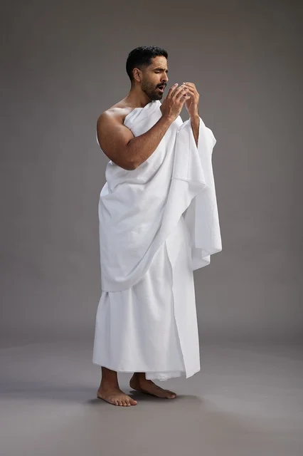 A portrait of a Saudi man in Ihram clothing sneezing and respectfully covering his mouth, the concept of Hajj and Umrah, full-body, gray background.