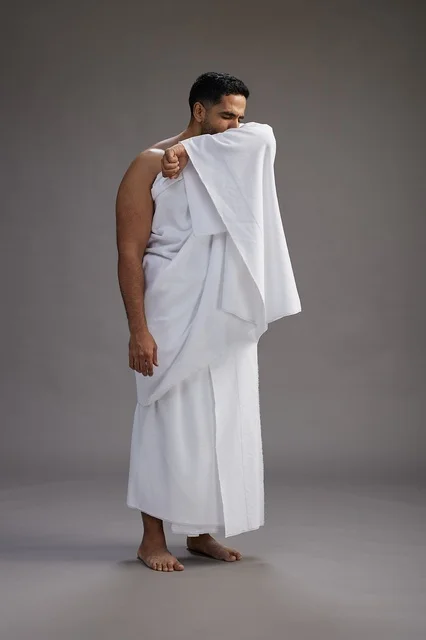 A portrait of a Saudi man in Ihram clothing sneezing and respectfully covering his mouth, the concept of Hajj and Umrah, full-body, gray background.