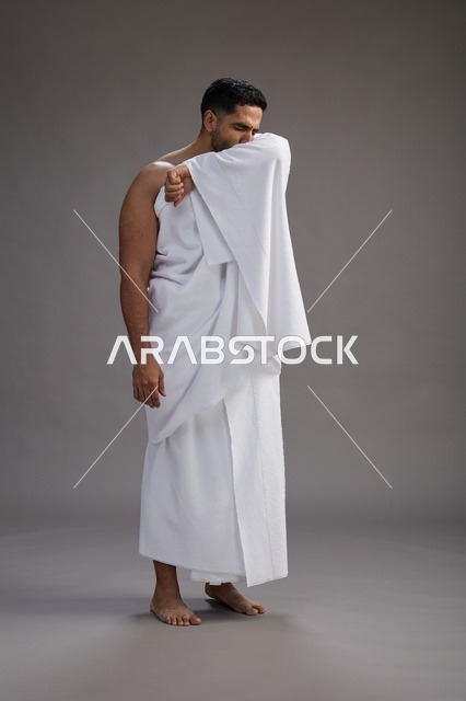 بورتريه لرجل سعودي بملابس الإحرام يعطس ويغطي فمه باحترام, مفهوم الحج والعمره, الجسم بالطول الكامل, خلفيه رماديه