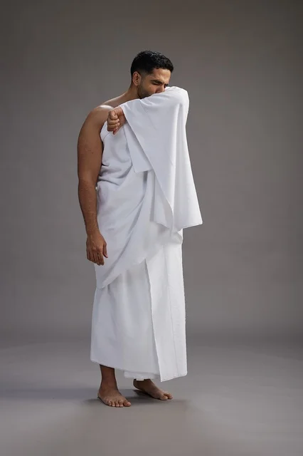 بورتريه لرجل سعودي بملابس الإحرام يعطس ويغطي فمه باحترام, مفهوم الحج والعمره, الجسم بالطول الكامل, خلفيه رماديه