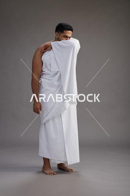بورتريه لرجل سعودي بملابس الإحرام يعطس ويغطي فمه باحترام, مفهوم الحج والعمره, الجسم بالطول الكامل, خلفيه رماديه