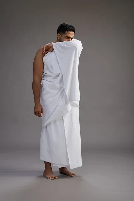 بورتريه لرجل سعودي بملابس الإحرام يعطس ويغطي فمه باحترام, مفهوم الحج والعمره, الجسم بالطول الكامل, خلفيه رماديه
