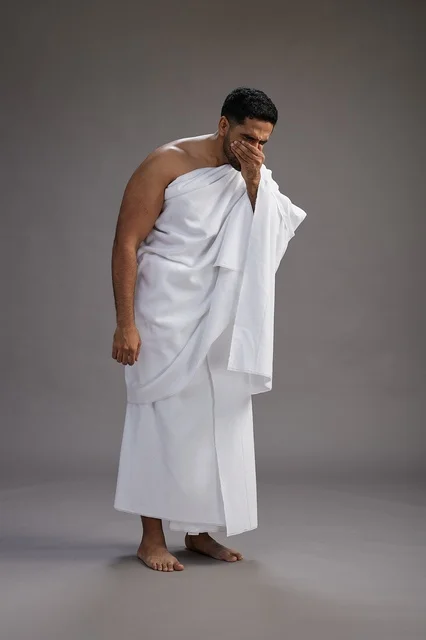 A portrait of a Saudi man in Ihram clothing sneezing and respectfully covering his mouth, the concept of Hajj and Umrah, full-body, gray background.