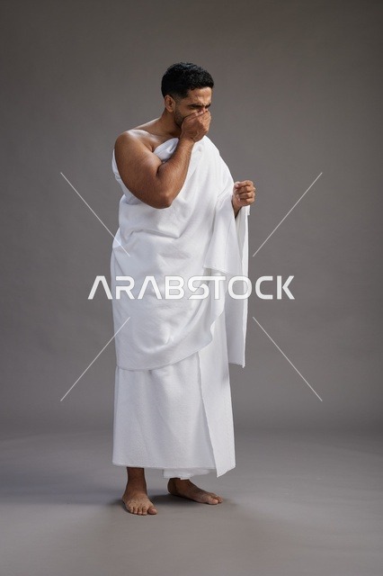 A portrait of a Saudi man in Ihram clothing sneezing and respectfully covering his mouth, the concept of Hajj and Umrah, full-body, gray background.