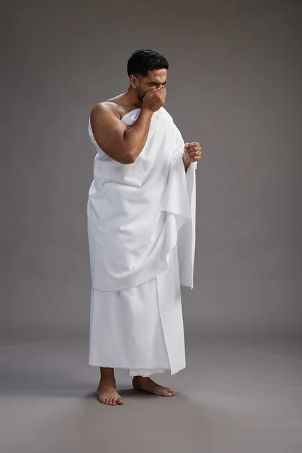 A portrait of a Saudi man in Ihram clothing sneezing and respectfully covering his mouth, the concept of Hajj and Umrah, full-body, gray background. A portrait of a Saudi man in Ihram clothing sneezing and respectfully covering his mouth, the concept of Hajj and Umrah, full-body, gray background.